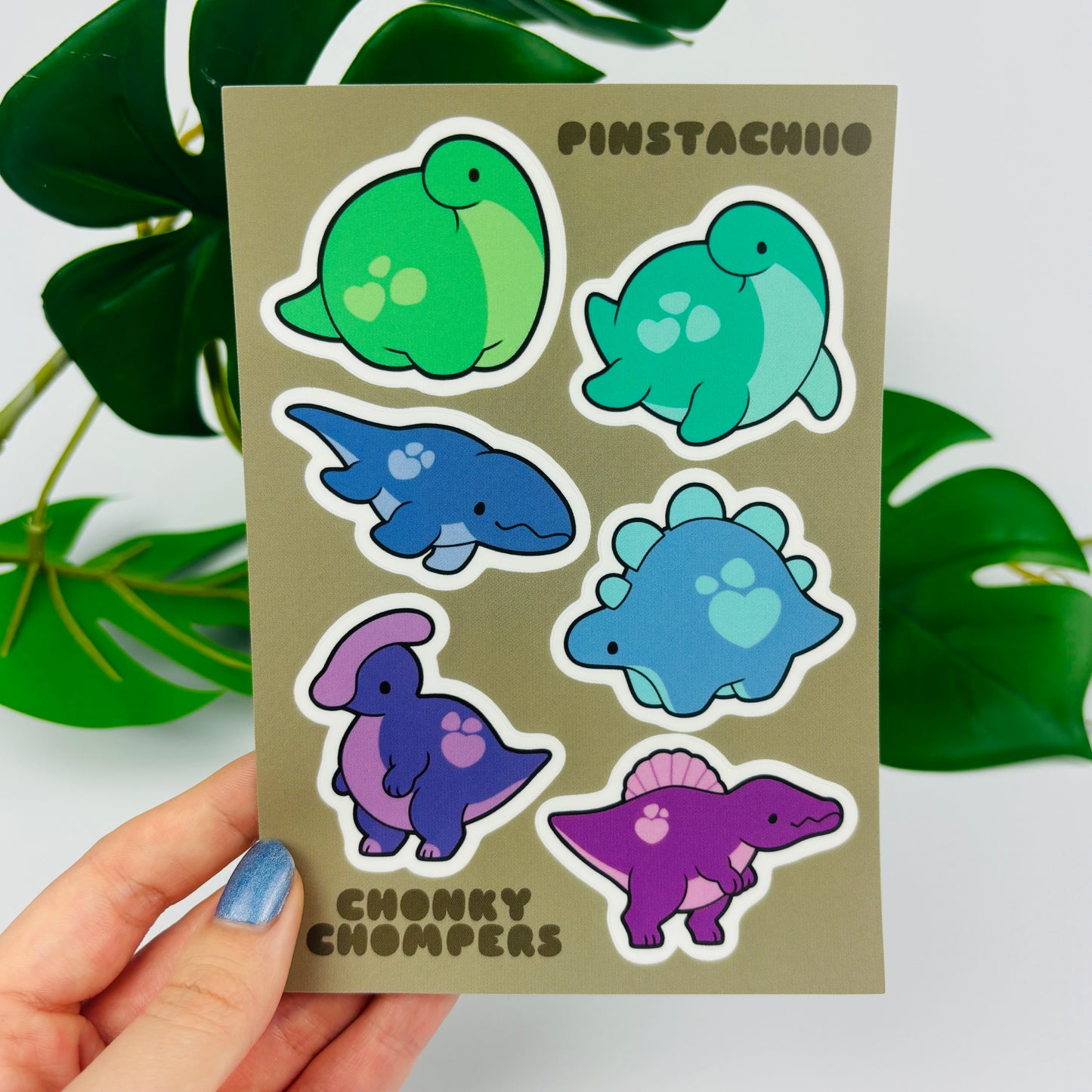 A hand holding a sheet of colorful dinosaur stickers with six different designs, including green, blue, and purple dinosaurs on a beige background.